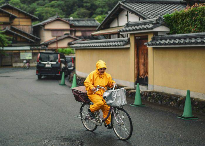 A woman in a Japanese raincoat, cycling along a traditional Japanese street in the rain.